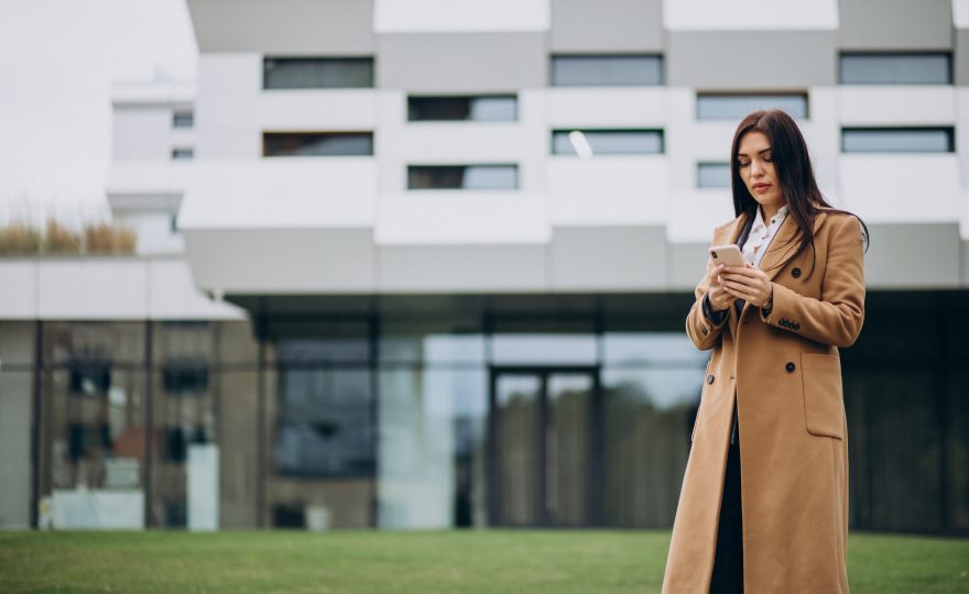 Mulher analisando informações no celular em frente a um prédio moderno, representando avaliação de investimentos em imóveis comerciais em Campo Grande.